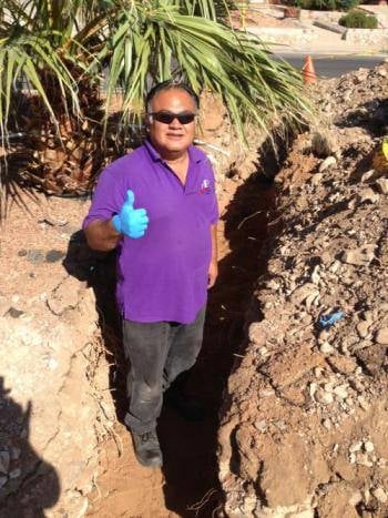 Person in purple shirt and sunglasses standing in sandy excavation pit with palm trees in background